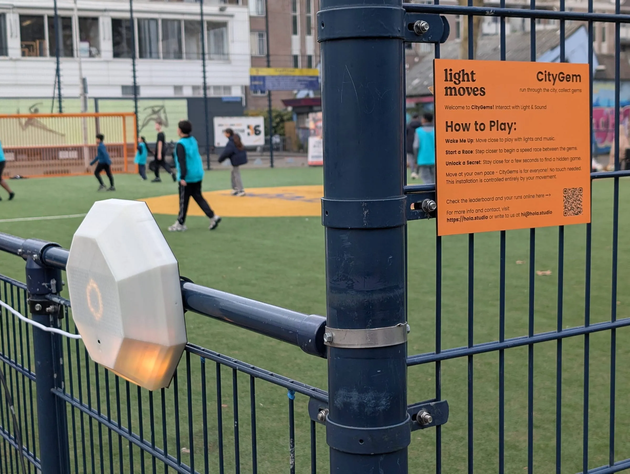 A CityGem module clamped to the fence of a Rotterdam sports court, lit, beside its how-to-play sign